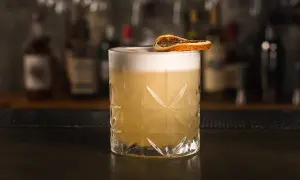 Klasyczny whiskey sour przepis: orzeźwiający koktajl z pianką i suszoną pomarańczą na tle barowych butelek.
