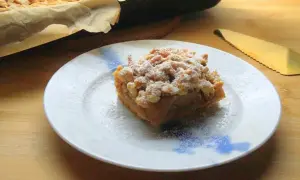 Szybka szarlotka z jabłkami ze słoika, posypana cukrem pudrem, na talerzu. Obok blacha z resztą ciasta i złota łopatka.