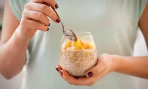 Ręce trzymają szklankę z deserem z chia i mlekiem kokosowym, ozdobionym kawałkami ananasa i sezamem.