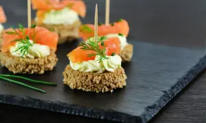 Eleganckie finger food: krążki ciemnego pieczywa z kremowym serkiem, łososiem i koperkiem, podane na łupkowej desce.