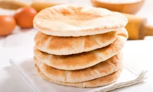 Stos świeżo upieczonego chleba arabskiego na białej serwetce, obok jajka i wałka do ciasta.