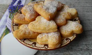 Stos ciasteczek serowych z 3 składników w kształcie serc, posypanych cukrem pudrem, na talerzu z lawendą w tle.