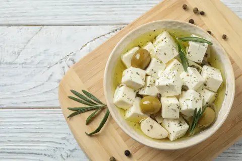 Kostki sera feta w oliwie z oliwkami, czosnkiem i rozmarynem, na desce.