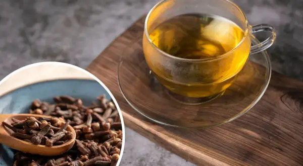 Aromatyczna woda z goździkami w szklanej filiżance, obok miseczka pełna goździków na drewnianej desce.