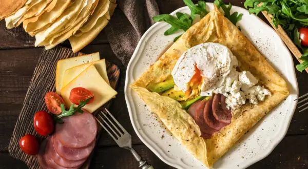Pyszne crepes z jajkiem w koszulce, awokado, szynką i serem. Obok stos naleśników i dodatki.