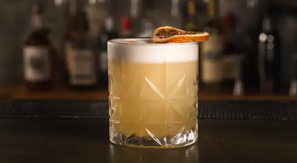 Klasyczny whiskey sour przepis: orzeźwiający koktajl z pianką i suszoną pomarańczą na tle barowych butelek.