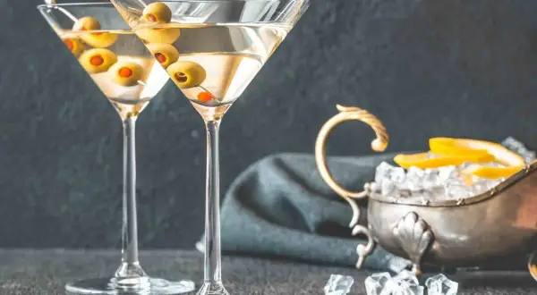 Dwa kieliszki martini z oliwkami, obok srebrna miseczka z lodem i cytryną. Idealne do podania z czym pić martini.