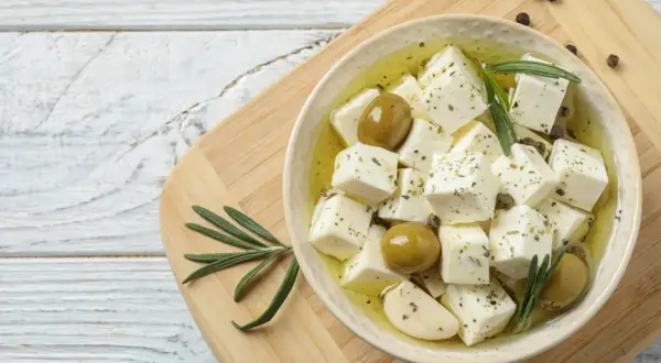 Kostki sera feta w oliwie z oliwkami, czosnkiem i rozmarynem, na desce.