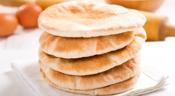 Stos świeżo upieczonego chleba arabskiego na białej serwetce, obok jajka i wałka do ciasta.