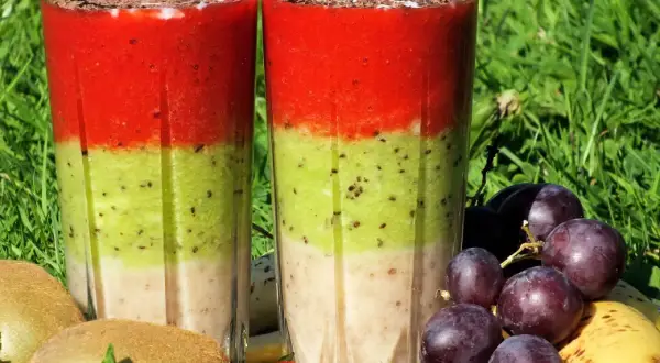 Warstwowy koktajl owocowy z truskawkami, kiwi i bananem, ozdobiony startą czekoladą. Obok owoce: winogrona, kiwi, truskawki, banan.