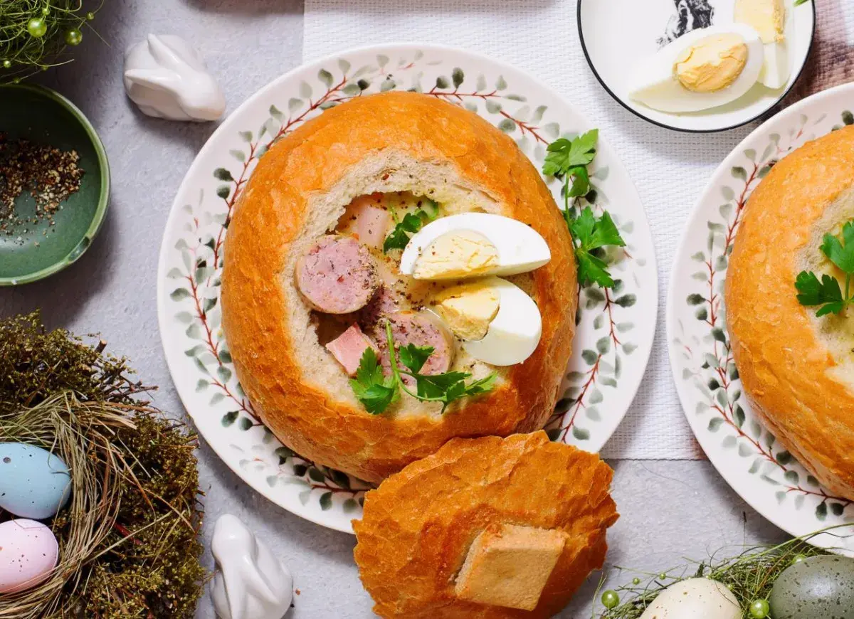 Pyszny żur w chlebie z kiełbasą i jajkiem, ozdobiony natką pietruszki. Na stole wielkanocne dekoracje: pisanki w gnieździe, zajączek.