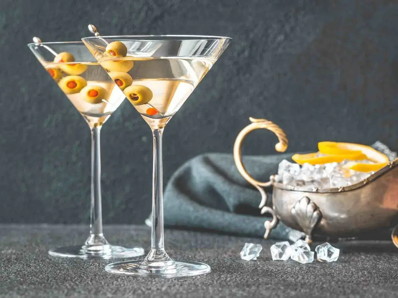 Dwa kieliszki martini z oliwkami, obok srebrna miseczka z lodem i cytryną. Idealne do podania z czym pić martini.