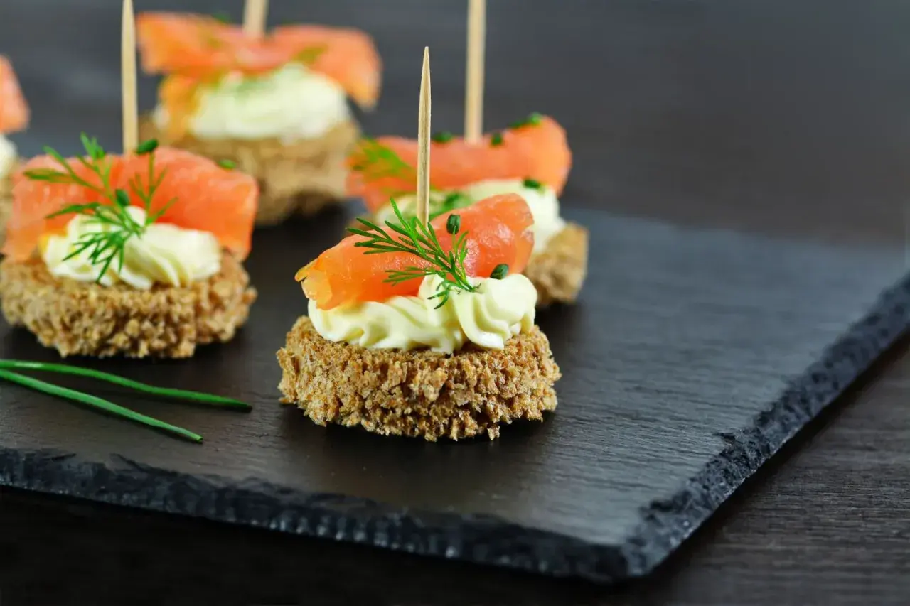 Eleganckie finger food: krążki ciemnego pieczywa z kremowym serkiem, łososiem i koperkiem, podane na łupkowej desce.