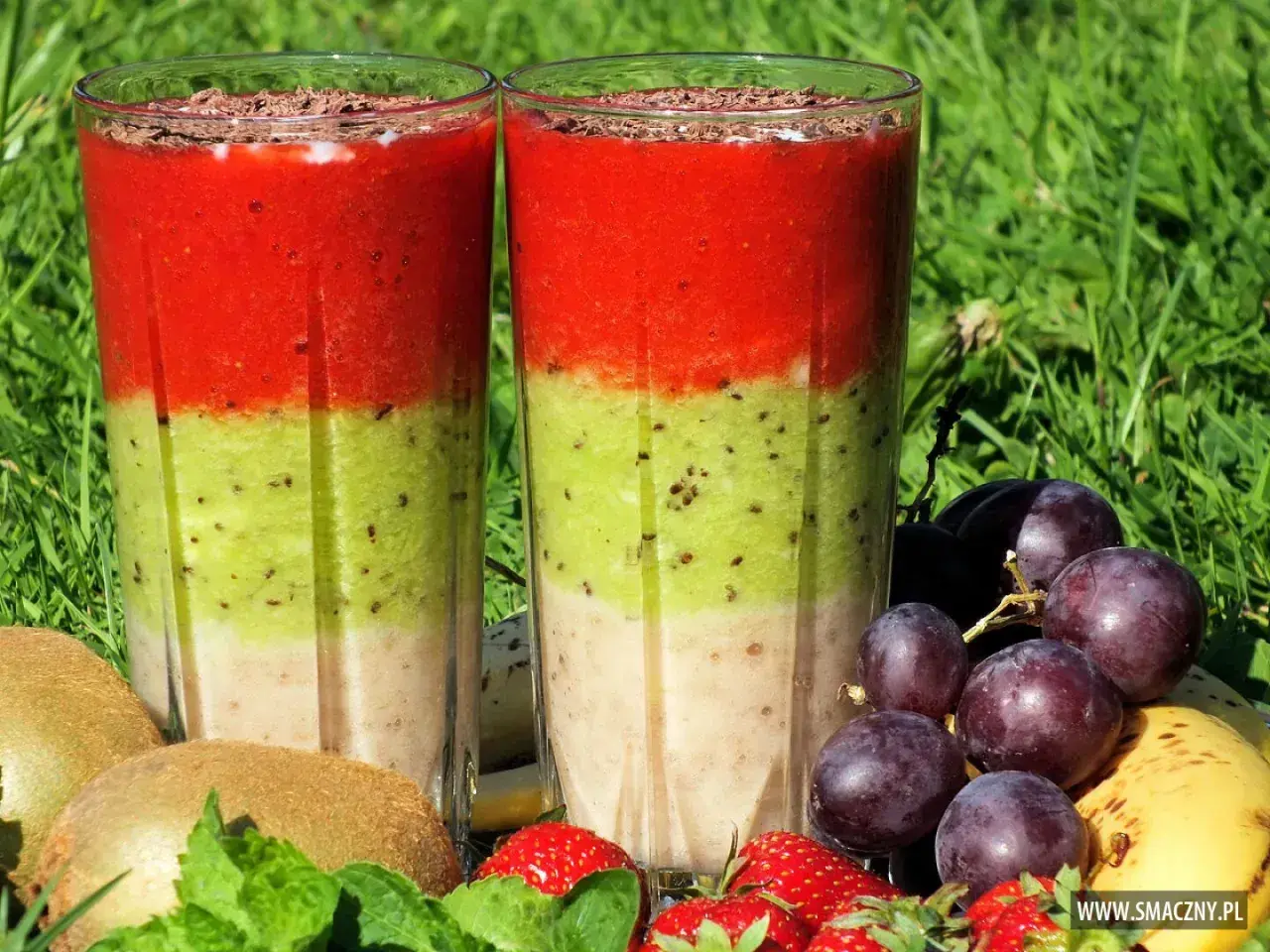 Warstwowy koktajl owocowy z truskawkami, kiwi i bananem, ozdobiony startą czekoladą. Obok owoce: winogrona, kiwi, truskawki, banan.