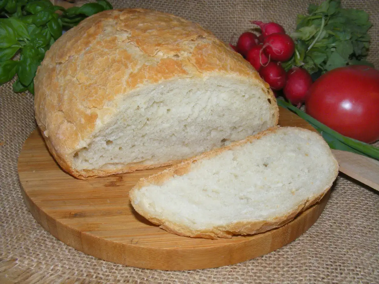 Pyszny, domowy chleb z garnka, pokrojony na desce, z pomidorem, rzodkiewkami i bazylią w tle.