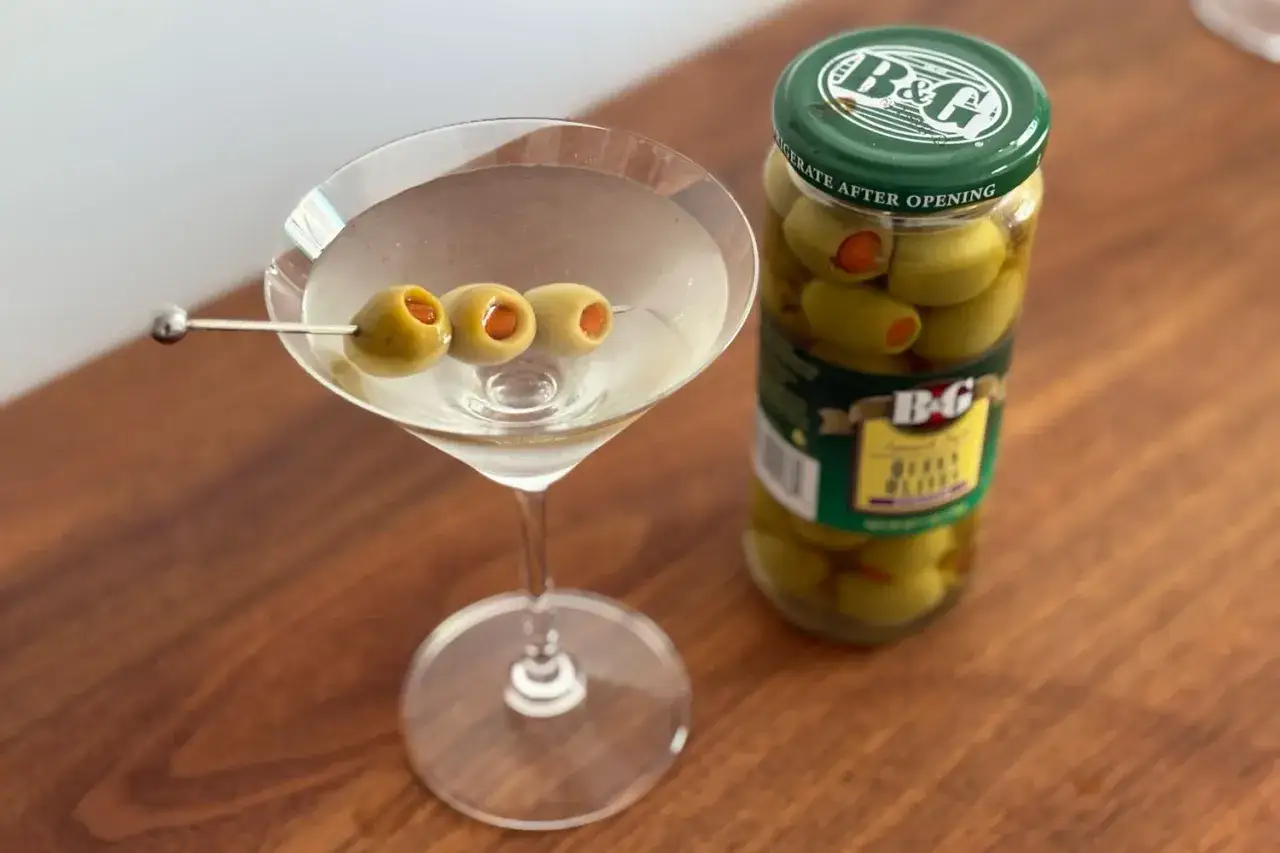 Idealne z czym pić martini? Z oliwkami! Kieliszek z drinkiem i szklany słoik pełen zielonych oliwek B&G na drewnianym blacie.