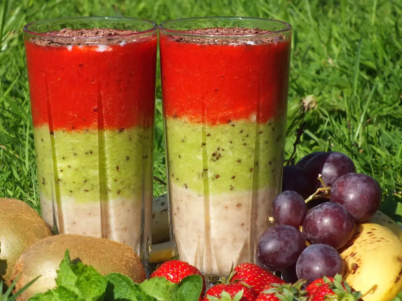 Dwa warstwowe koktajle owocowe: truskawkowy, kiwi i bananowy, ozdobione startą czekoladą. Obok świeże owoce: kiwi, winogrona, truskawki i banan.