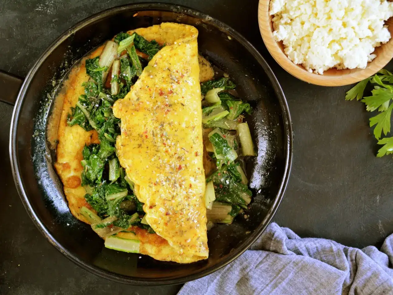 Puszysty omlet z serka wiejskiego z zielonymi warzywami na patelni. Obok miseczka z serkiem i natka pietruszki.