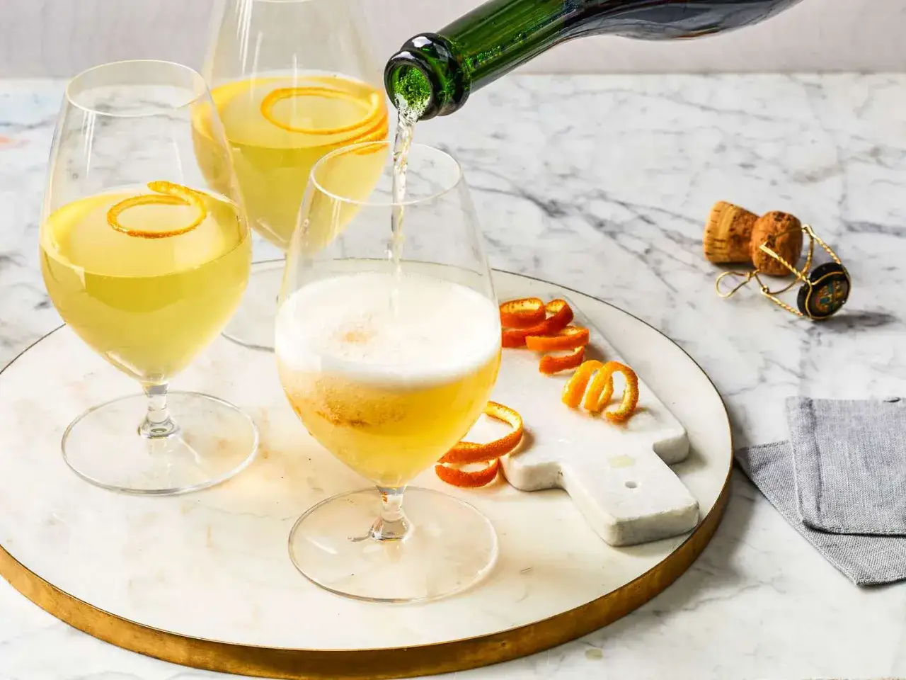 Trzy kieliszki musującego napoju, jeden nalewany z butelki. Każdy ozdobiony skórką pomarańczy. Idealna mimosa na brunch.