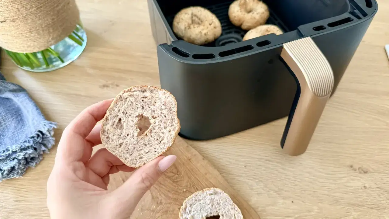 Keto pieczywo w air fryerze. Ręka trzyma przekrojony bagelek, w tle urządzenie z innymi wypiekami.