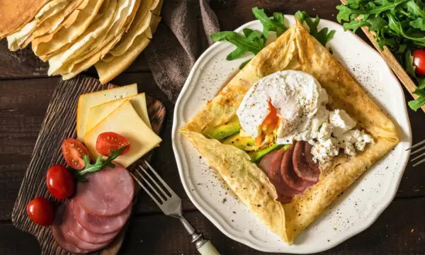 Pyszne crepes z jajkiem w koszulce, awokado, szynką i serem. Obok stos naleśników i dodatki.