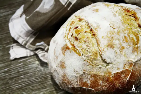 Świeżo upieczony bochenek chleba na drożdżach, oprószony mąką, leży obok lnianej ściereczki. Idealny przepis na chleb na drożdżach.