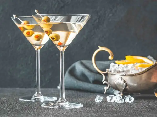 Dwa kieliszki martini z oliwkami, obok srebrna miseczka z lodem i cytryną. Idealne do podania z czym pić martini.