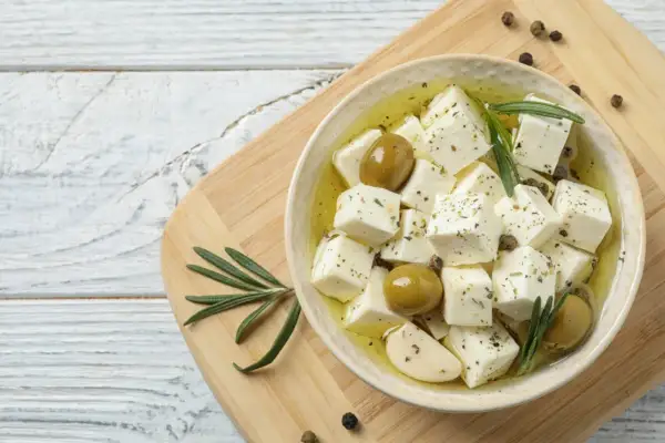 Kostki sera feta w oliwie z oliwkami, czosnkiem i rozmarynem, na desce.