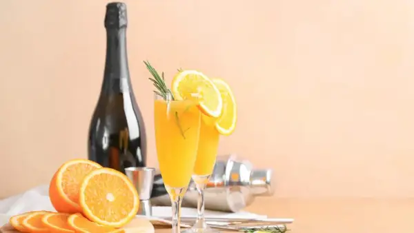Dwa kieliszki orzeźwiającej mimosa z plasterkami pomarańczy i gałązką rozmarynu, obok butelki szampana i shakera.